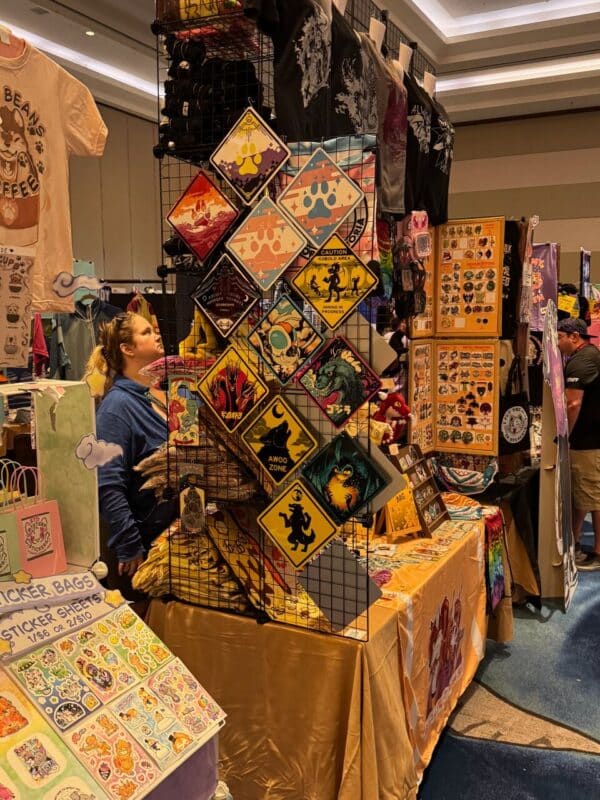 A convention booth displaying a variety of colorful and whimsical merchandise. The booth features t-shirts, vibrant art prints, and signs with paw print designs and humorous slogans like “AWOO ZONE.” There are sticker sheets on the left, with playful, cartoonish designs. A shelf displays more stickers and art prints, while the table is draped with a