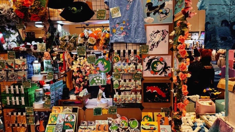 A vibrant market stall displays an array of colorful items. On display are art prints featuring whimsical creatures, alongside clothing items like a blue t-shirt adorned with stars. Various accessories and trinkets, such as pins and patches, are creatively arranged. The stall is decorated with floral garlands, adding to the lively, festive atmosphere. Prices are visible on
