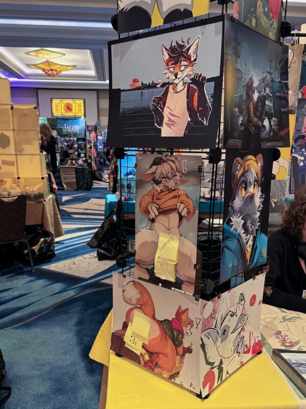 A display at a convention showcasing various pieces of anthropomorphic art. The art features animal characters with human-like features in colorful, dynamic poses. Some artworks have sticky notes with prices or messages like Goated. In the background, attendees browse other stalls with merchandise visible. The setting is well-lit with a ceiling featuring decorative lights, and the floor is