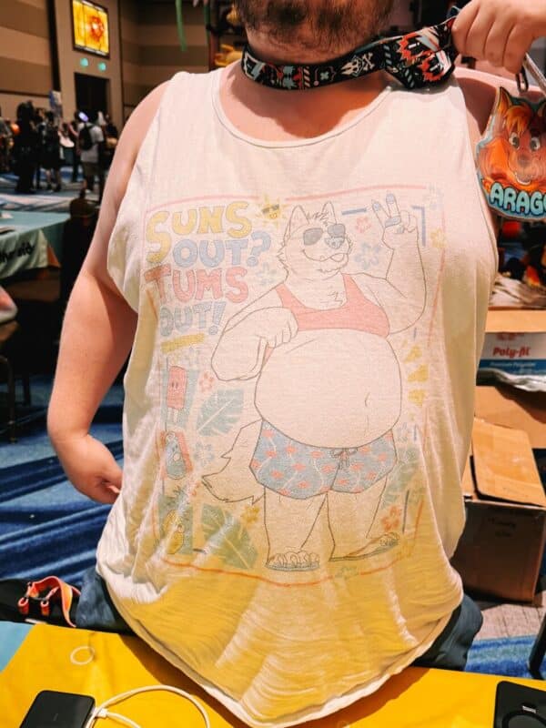 A person wearing a white tank top featuring a cartoon illustration of an anthropomorphic dog dressed in summer attire, with sunglasses and a peace sign gesture. The shirt has the text SUNS OUT? TUMS OUT! in colorful lettering. The individual is holding a decorated lanyard with a badge featuring a fox illustration clipped to it. The background