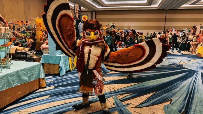 A person is wearing an elaborate owl costume at a convention. The costume features large, detailed wings with a brown, white, and red pattern, resembling feathers. The owl head has expressive eyes and a yellow beak. The person stands with their wings spread, showcasing the intricate design. The background shows a busy convention hall with various tables covered in light