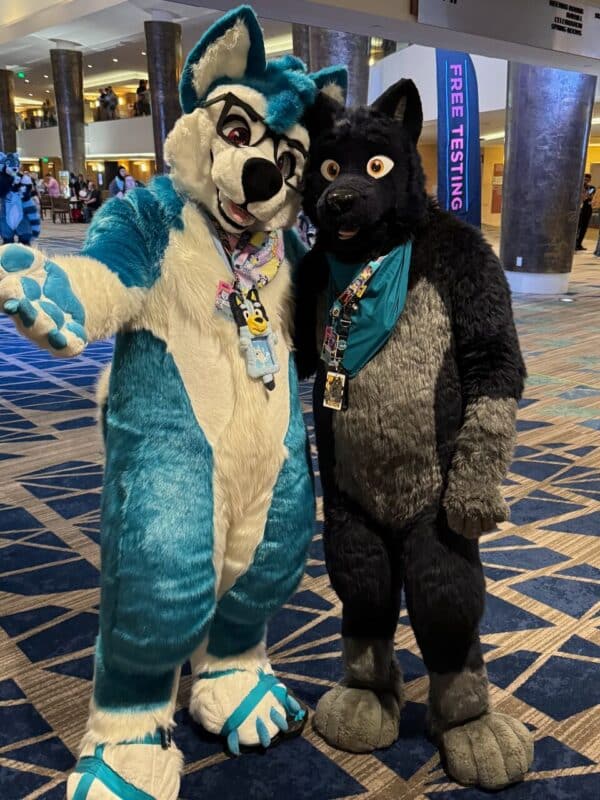 Two people in colorful fursuits are posing in a convention center. The left person is wearing a teal and white suit with glasses and has a playful expression. The right person is in a black and gray suit with bright eyes. Both are wearing multiple badges and lanyards. The background shows other attendees and a sign that reads Free Testing.
