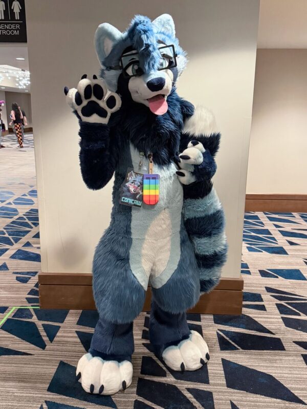 A person in a blue and white raccoon-like furry costume, complete with glasses and a wagging tongue, stands indoors on a patterned carpet, waving with one paw. They have white and black paw pads and a large bushy tail. Around their neck, they wear a badge featuring a colorful pride flag. In the background, a partial view of