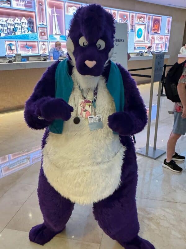 A person in a purple and white furry costume stands in a brightly lit hotel lobby. The costume features a cartoonish animal character with a blue vest and a large fabric tail. The person has a lanyard around their neck displaying a badge. In the background, there's a decorated wall with small artworks, and another person is visible behind a counter,