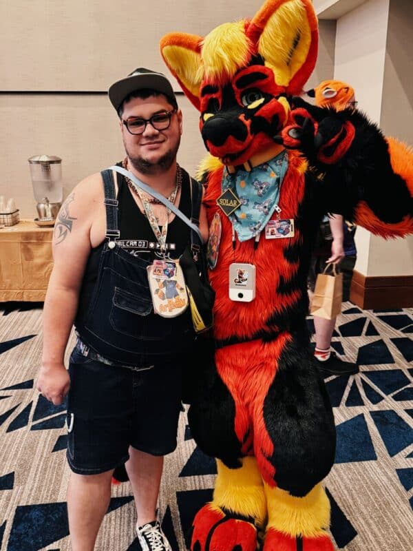 A person in a black tank top, shorts, and cap stands next to someone wearing an elaborate red, black, and yellow furry costume, resembling a fox with large ears. The costumed individual has a colorful bandana around the neck and a badge with the name Solar. Both have lanyards with various badges. Behind them, a table