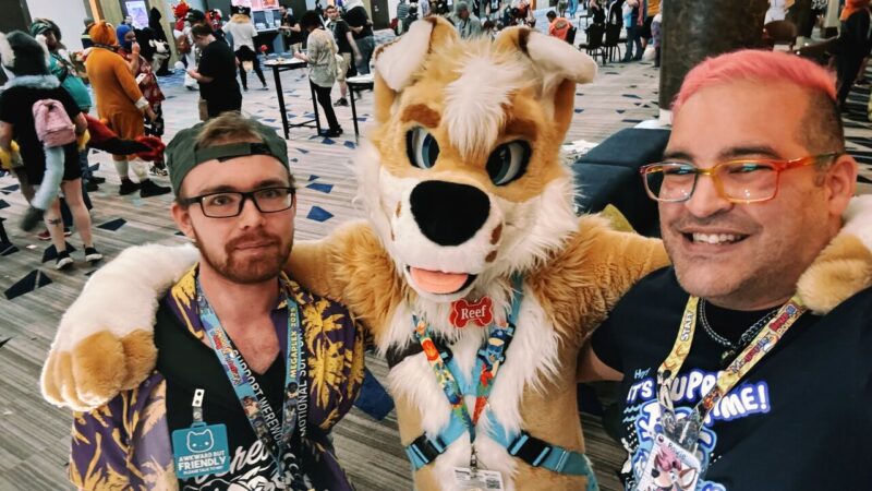 Two people stand on either side of a person in a light brown and white furry costume with large blue eyes, sticking out a red tongue, and wearing a name tag labeled Reef. The person on the left wears glasses, a patterned shirt, and a cap, displaying a badge reading Awkward But Friendly. The person on the right