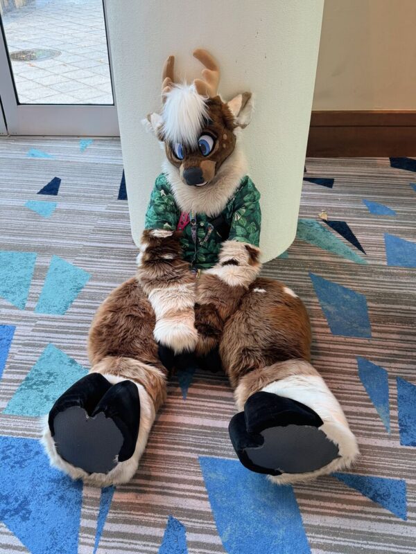 A person in a deer fursuit is sitting on a patterned carpet floor, leaning against a white pillar. The fursuit features brown fur, light tan antlers, and large blue eyes, with a tuft of white hair on the head. The suit is wearing a green shirt with a leafy design and badges on the chest. The floor has
