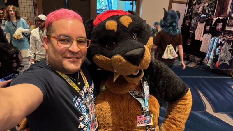 A person with pink hair and glasses smiles for a selfie with someone in a large dog-like fursuit, featuring black and tan fur, along with a red hat. Both wear lanyards with badges. In the background, there are several other people, including individuals in costumes, walking around a convention-style setting with vendor booths displaying various items like