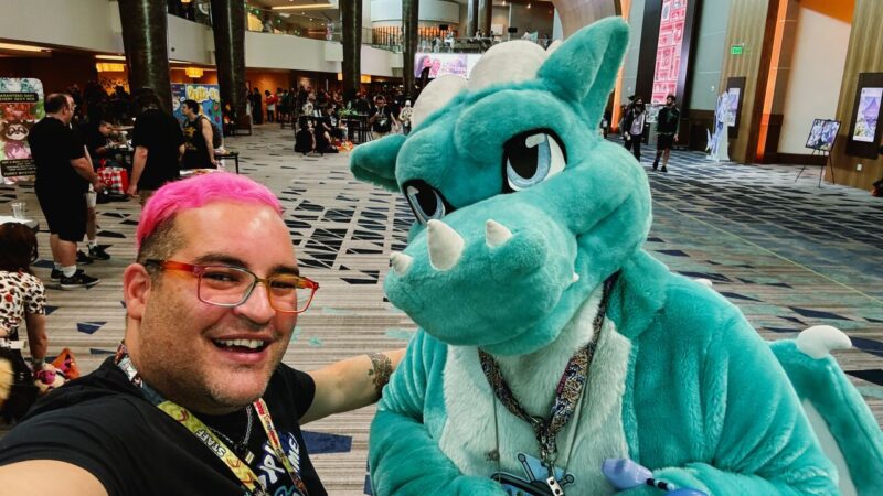 A person with pink hair and glasses takes a selfie with a person in a turquoise dragon costume. They are both smiling and wearing badges around their necks. The background shows a spacious convention hall with people mingling, colorful displays, and posters. The atmosphere is lively and festive, suggesting a pop culture or comic convention event.
