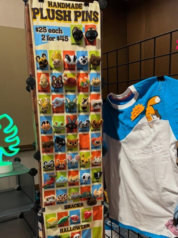 A display of handmade plush pins is shown. Each pin features an animal face, with options like fox, bat, panda, and more. The sign reads $25 each, 2 for $45. The display is colorful, with each pin housed in its own square compartment. Next to the display is a blue and white shirt with a cartoon fox
