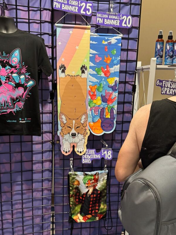 A merchandise display at a convention features colorful goods. On the left is a black T-shirt with a vibrant, neon design of a cat. In the center, two pin banners are on display: one with an illustration of a corgi and a pastel rainbow background, priced at $25, and another depicting colorful balloon animals against a blue sky, priced