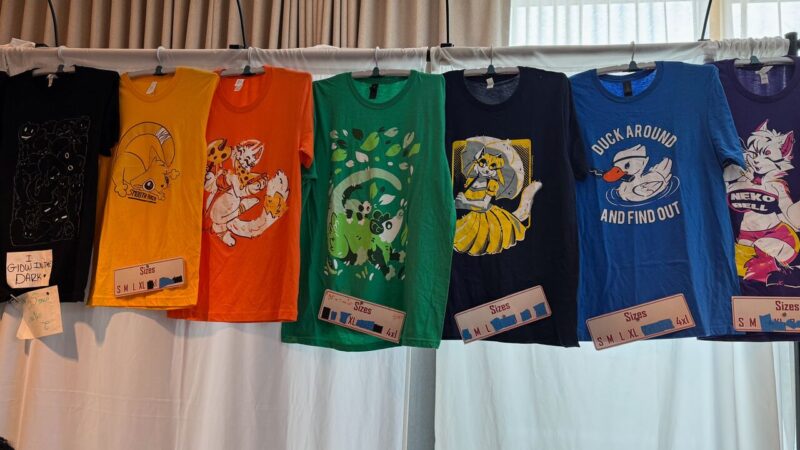 A row of colorful T-shirts is displayed hanging on a rack. The designs feature various illustrations, including a black shirt with glow-in-the-dark figures, a yellow shirt with a character inside a swirl, an orange shirt with a cartoon fox holding mushrooms, a green shirt with a leafy pattern and animals, a navy shirt with a character in a yellow dress