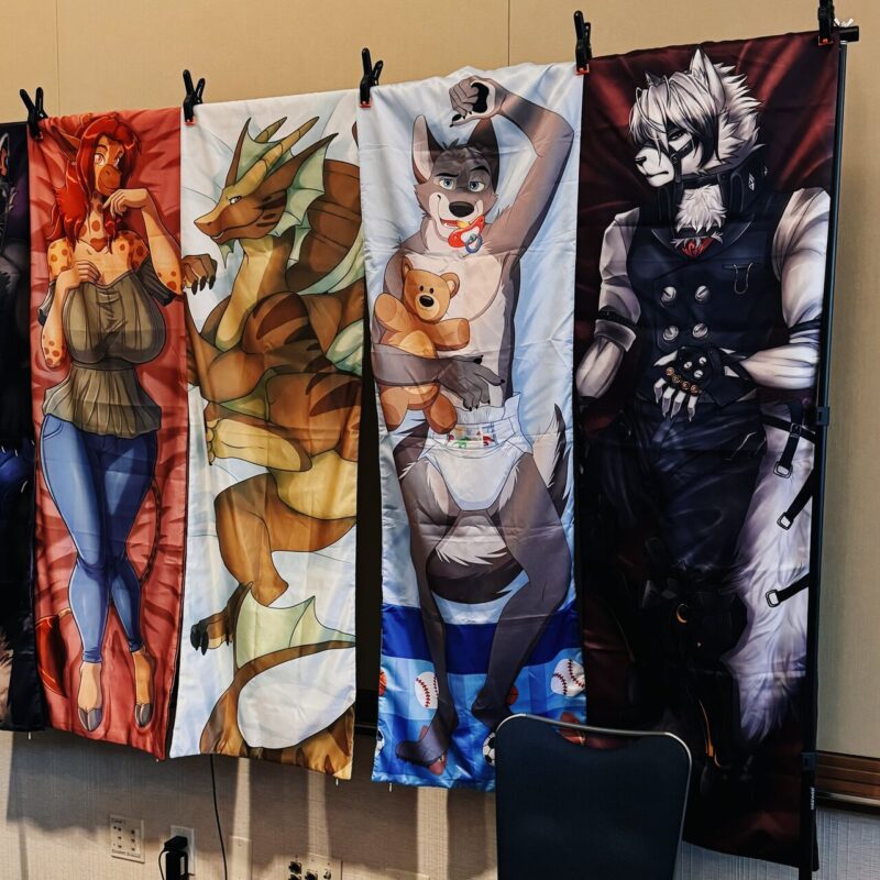 Four colorful banners hang side by side, each depicting an anthropomorphic character. From left to right: a red-haired character with a tail in a sweater and jeans; a dragon-like creature with horns and wings; a canine character wearing a diaper and holding a teddy bear; a white-haired wolf in dark, detailed clothing with gloves and straps. The banners