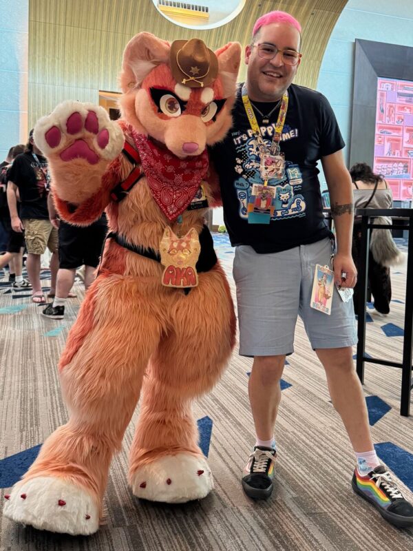 A person stands next to someone in a detailed orange and cream anthropomorphic fox costume, which features a cowboy hat, a red bandana, and large paw gloves. The costumed individual is posing playfully with one paw raised. The person beside them has pink hair and glasses, wearing a black shirt with white and blue graphics, light gray shorts,