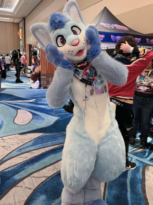 A person is wearing a light blue and white furry costume with a large, cartoonish animal head featuring big eyes and blue tuft of hair. They're posing playfully with their hands on their cheeks. The costume has various badges attached to it. In the background, other people are present, some in costumes, and there's a booth with Weasel Con