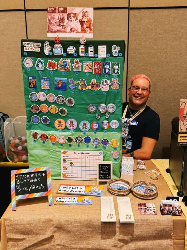 A person with pink hair and glasses smiles while sitting behind a display table covered with a variety of colorful stickers and buttons featuring cartoon characters and humorous text. The display board is green with several rows of circular pins and stickers. The table also has small baskets and promotional materials, including signs saying Stickers + Buttons and Who Let a Baby Drive