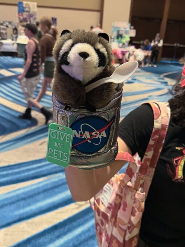 A plush raccoon toy is nestled inside a silver cup with a NASA logo. The raccoon holds a white plastic spoon. Attached to the cup is a green tag reading GIVE ME PETS. The background shows an indoor setting with people and tables, perhaps a convention or event, and patterned blue carpeting. Part of a person’s arm holding