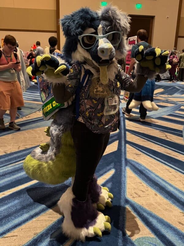 A person is wearing a colorful, fun costume with vibrant green, gray, and white fur. The outfit features exaggerated features like large paws and a snout. They are accessorized with oversized sunglasses and a patterned shirt complete with a name badge. The person is playfully posing with their paws raised. In the background, other people are walking around a