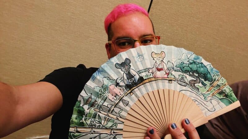 A person with pink hair and glasses holds a decorative fan partially in front of their face. The fan features an illustration of two anthropomorphic mice in traditional clothing, sitting calmly in a serene landscape with trees and mountains. The person is wearing a black shirt and has their nails painted in metallic colors. The background is a plain, neutral-colored wall.