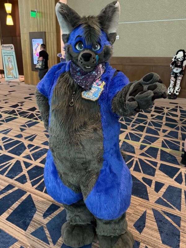 A person dressed in a vibrant blue and gray furry costume with large ears and playful eyes stands on a patterned carpet in a hallway. They have a friendly, inviting pose with one hand extended forward and wear a starry bandana around their neck. A colorful name tag is attached to the costume. In the background, there’s signage indicating Megap