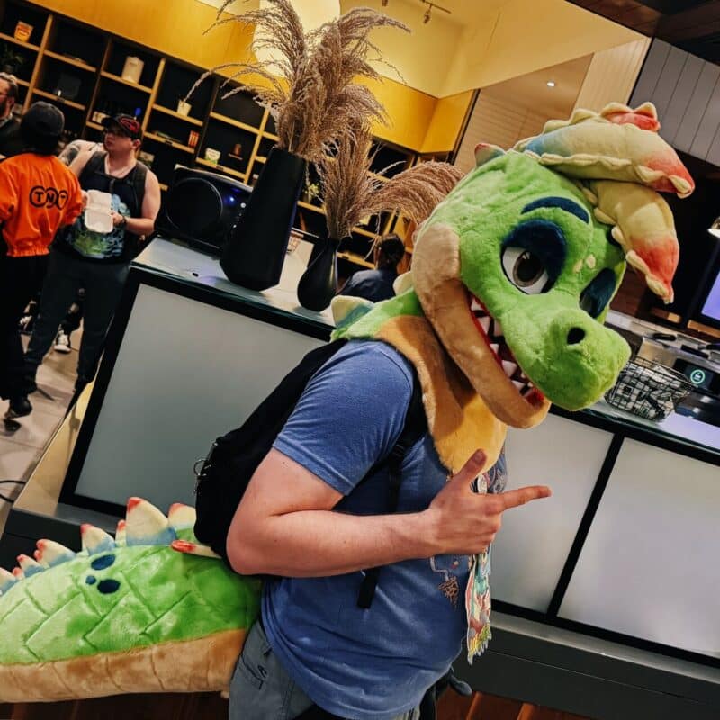 A person wearing a colorful green dinosaur costume poses indoors. The costume features a playful expression with large eyes, a wide smile, and a rainbow mane. The person is wearing a blue shirt and a black backpack, giving a peace sign with one hand. The background shows a modern room with shelves, people, and vases holding decorative grasses. Others in