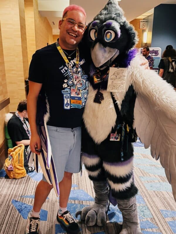 A person with pink hair and glasses smiles while standing with an individual in a detailed bird-like fursuit. The fursuit features large wings, a beak, and vibrant purple, black, and white colors. Both wear convention badges and lanyards. The person on the left is wearing a black t-shirt and shorts, and they appear