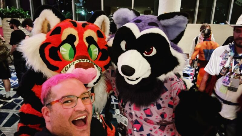 A person with pink hair and glasses is smiling widely while taking a selfie with two people in brightly colored animal costumes. The first costume is a red and black cat with green eyes, and the second is a purple and black animal with ears and a white snout. The background shows other people in costumes and lanyards, inside a large room with