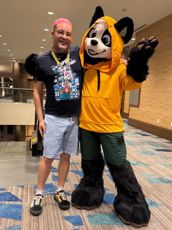 A person with pink hair, glasses, and a blue graphic T-shirt with light blue shorts stands smiling beside someone in a large, cartoonish panda costume. The panda suit features a vibrant orange hoodie and green shorts, with black and white fur accents. The two pose together in a well-lit indoor setting with modern design elements like blue and brown geometric carpet