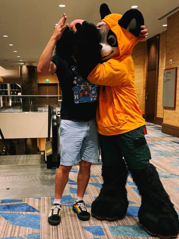 A person wearing casual clothes with a colorful sneaker design is playfully interacting with someone in a furry panda costume. The panda costume features a hooded orange shirt and large black paws. They are standing on a carpeted floor that has blue and gray patterns, in what appears to be a hotel lobby or convention center setting. The scene is well-lit,