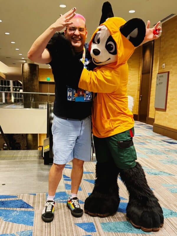 A person with pink hair smiling and throwing a hand sign stands next to a person in a panda costume. The costume features a large white and black panda head, orange hoodie, and green shorts. They are posed with arms around each other in a playful manner. The scene is indoors on a patterned floor in a well-lit hallway, possibly at a convention
