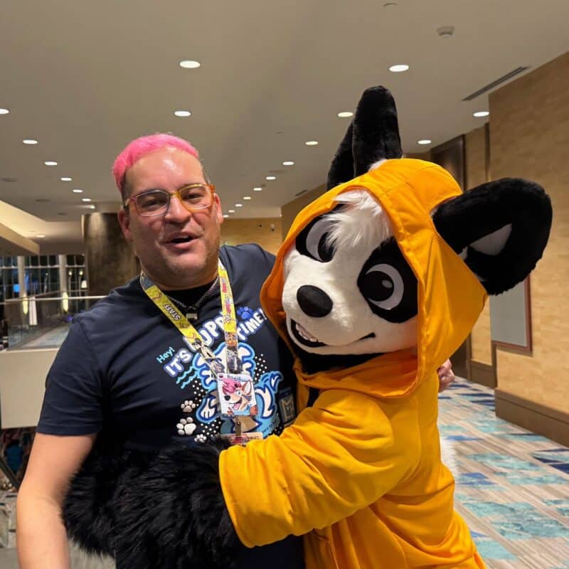 A person with pink hair and glasses is smiling and holding a plush costume character. The person is wearing a dark blue T-shirt and has a lanyard around their neck. The character resembles a panda or raccoon with black and white fur and is wearing a bright orange hoodie. They are indoors, in a well-lit hallway with modern decor and a