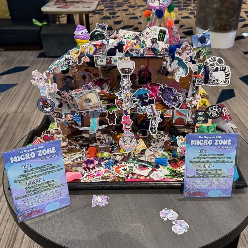 A colorful tabletop display titled The Megaplex 2023 Micro Zone features an array of small stickers, plushies, and decorative items. The display is vibrant, with cartoon characters and various small objects densely arranged. Signs explain rules for interaction: DO take a few items and DO NOT remove the structure. The scene is lively