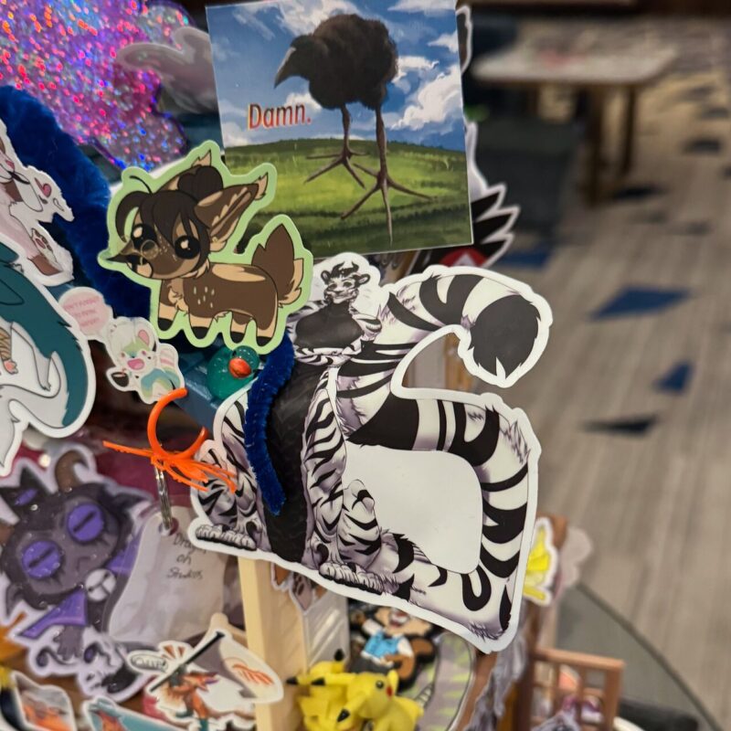 A collection of colorful stickers is displayed on a surface. The stickers include a cartoon deer with large eyes, a stylized black-and-white striped creature resembling a zebra, and a humorous illustration of a bird with the text “Damn.” There's also a 3D pop of embellishments like a blue fuzzy pipe cleaner and an orange plastic piece. In