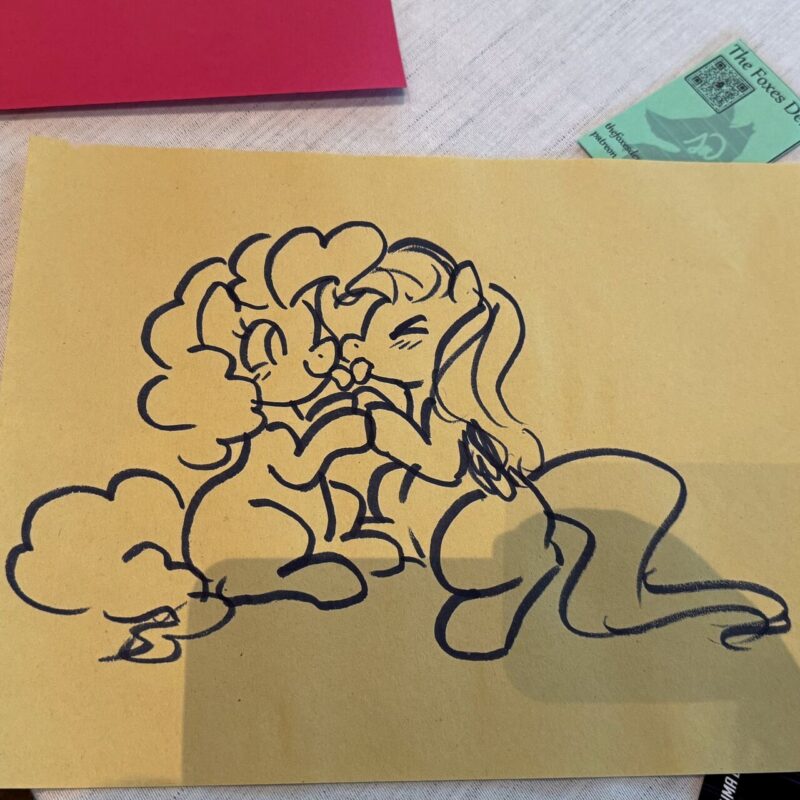 A drawing on yellow paper depicts two cartoon ponies sitting close together, smiling and holding hooves. Both ponies have distinct manes; one is curly, and the other is wavy. The drawing is surrounded by a table surface with a red paper and a small green-colored book visible in the background, placed on a light fabric.