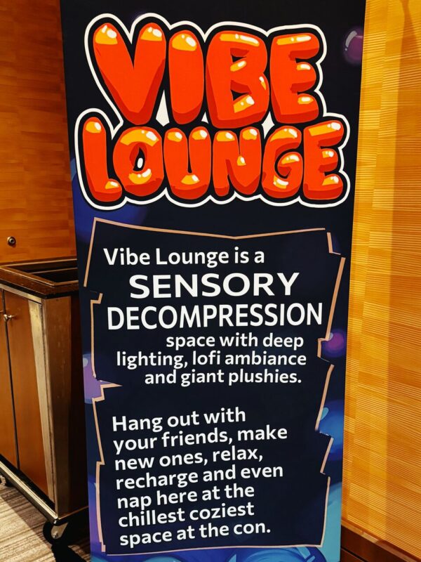 A vertical sign in a dimly lit area reads: Vibe Lounge in large, playful orange lettering at the top. Below, it describes the lounge as a SENSORY DECOMPRESSION space with deep lighting, lofi ambiance, and giant plushies. The text invites attendees to Hang out with your friends, make