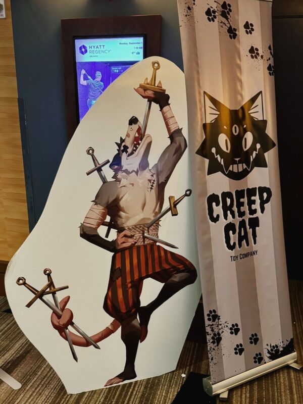 Cutout of an anthropomorphic wolf character performing a balancing act with swords, standing on one leg. The character wears bandages and striped pants, holding swords with its hands and tail. Beside it, a banner reads Creep Cat Toy Company with a stylized three-eyed cat logo and paw prints. In the background, a screen displays