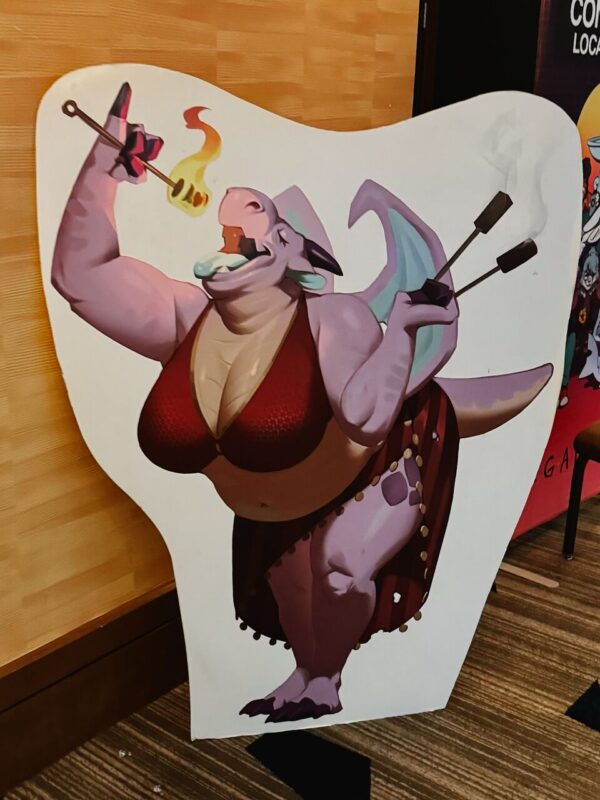 A whimsical cutout of a purple dragon character is displayed against a wooden wall. The dragon is anthropomorphic, wearing a red-scaled bikini top and belly dancer skirt adorned with gold coins. It is joyfully breathing fire toward a stick with burning marshmallows, holding an empty skewer in the other hand. The cutout has a playful and