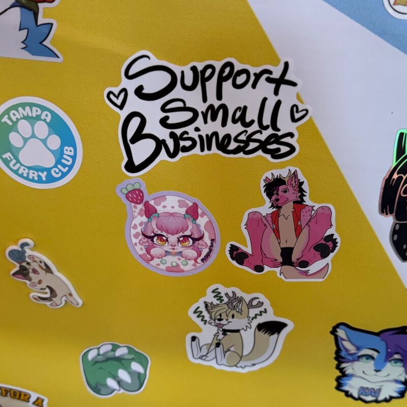 The image shows a collection of colorful stickers on a yellow background. In the center, a prominent sticker reads Support Small Businesses in playful, black script with heart accents. Surrounding it are various illustrated stickers, including cartoon characters and animals, such as a pink, horned character, a cute creature with pink curly hair and large eyes, and
