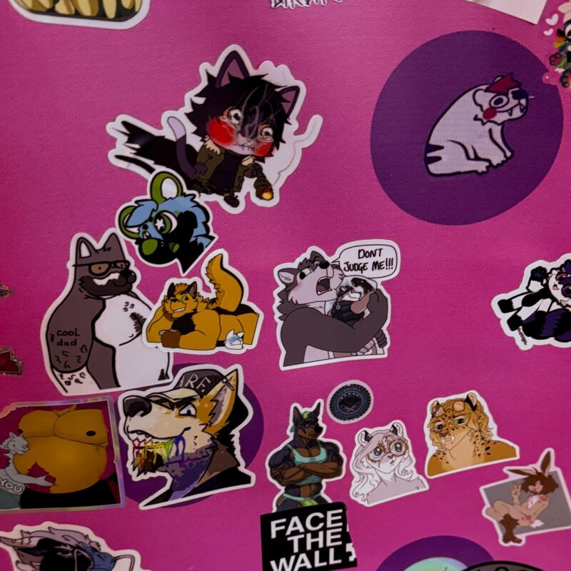 A collection of colorful stickers on a pink background featuring various cartoon animals and anthropomorphic characters. The illustrations include a blushing wolf-like character, a dog with glasses labeled “cool dad,” and a raccoon holding a smaller character with a speech bubble saying, “DON’T JUDGE ME!!!” There's a cat sitting on an oval, a wolf with