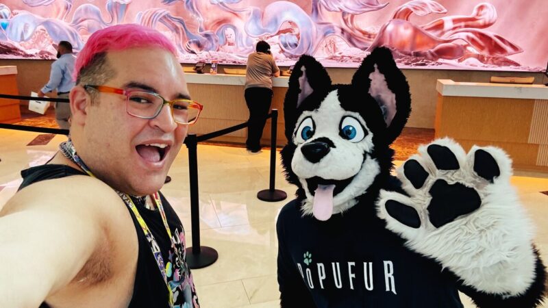 A person with pink hair and rainbow glasses takes a selfie with a friend in a black and white husky fursuit. The husky costume features bright blue eyes and an open mouth with its tongue out. The person smiles widely, wearing a black top and a colorful lanyard. Behind them, two people are at a counter, with an