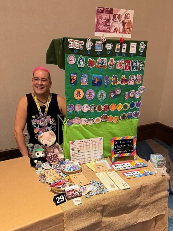 A person with pink hair sits behind a vendor table displaying various colorful merchandise, including stickers, buttons, and illustrated cards. The backdrop features an array of decorative items with animal designs, vibrant characters, and playful slogans. Plush toys and decorative items sit on the table, alongside signs advertising pricing, such as “Stickers $3 & Buttons 2/$