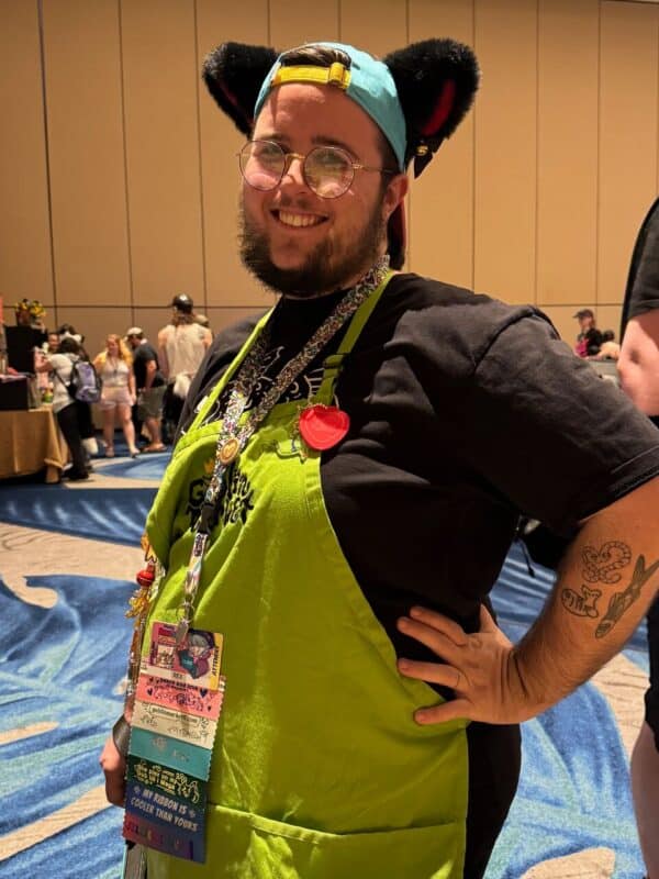 A person is smiling brightly at a convention. They wear a light blue cap with dark fuzzy ears, round glasses, and a bright green apron covered with various colorful ribbons and pins. Their arm is decorated with tattoos of a snake, fish, and plant. In the background, people are gathered, suggesting a busy, social event. The person’s confident