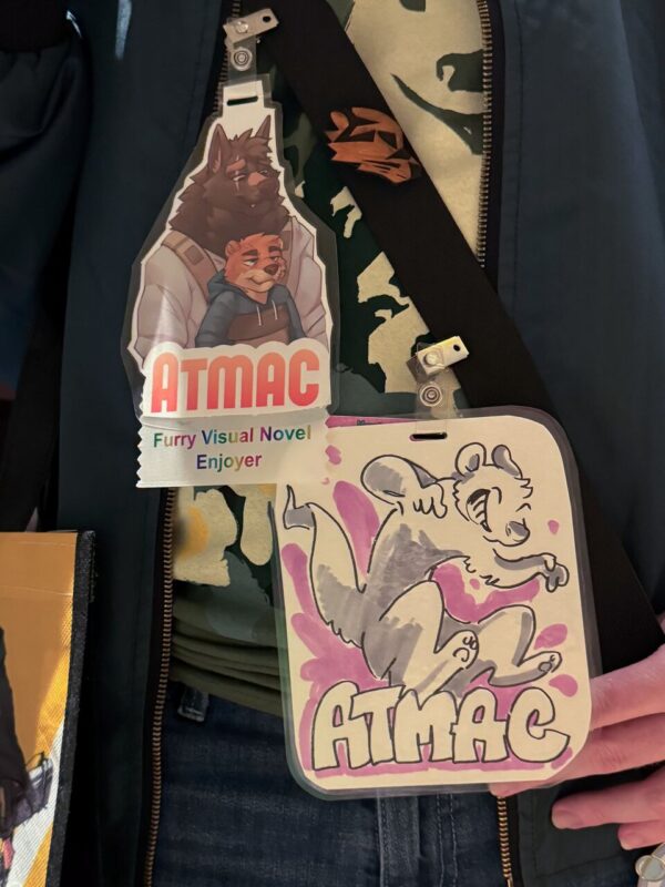 A person is wearing two badges around their neck. The first badge depicts two anthropomorphic animals, a wolf and a tiger, above the words ATMAC in bold red letters, with Furry Visual Novel Enjoyer written below. The second badge features a playful illustration of a leaping furry creature, surrounded by pink splashes, with