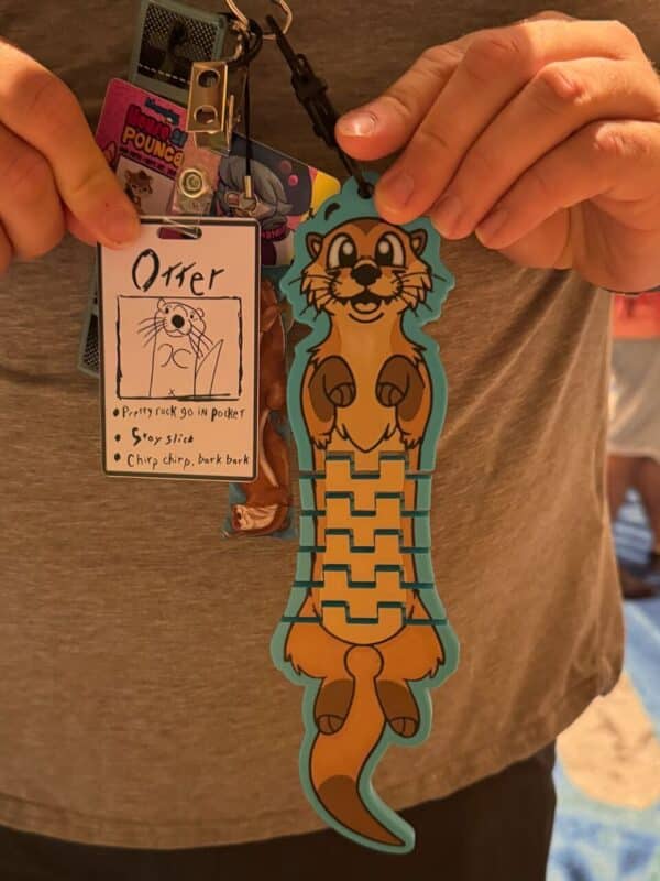 A person is holding a collection of badges and tags. One card features a simple drawing and text reading Otter, followed by humorous descriptions: Pretty rock go in pocket, Stay slide, and Chirp chirp, bark bark. Another tag displays a cartoon otter with a teal border and a playful expression. The person is wearing