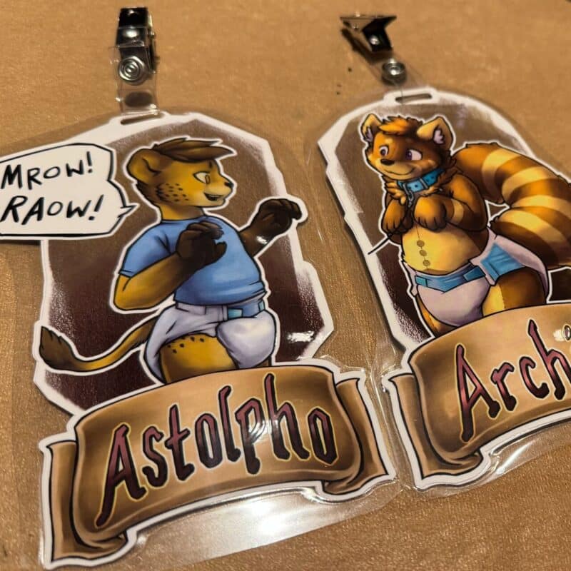 Two laminated badges featuring cartoon animal characters. The left badge depicts a yellow lion wearing a blue shirt and diaper, with speech bubbles saying Mrow! and Raow! It is labeled Astolpho. The right badge shows a raccoon-like creature with a striped tail, also in a diaper, with a shy expression, labeled Arch
