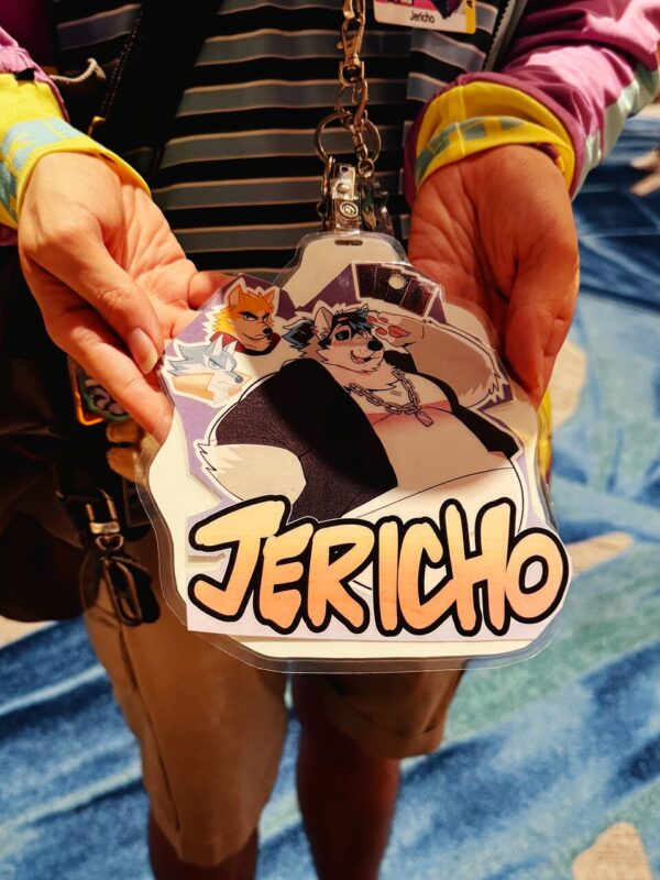 A person wearing a colorful striped and patterned shirt holds a large, laminated badge with cartoon-like drawings of anthropomorphic animals, including a wolf and a fox. The badge prominently features the name Jericho in bold, stylized letters. The background includes a blurry, abstract blue pattern on what appears to be a carpeted floor. The person’s