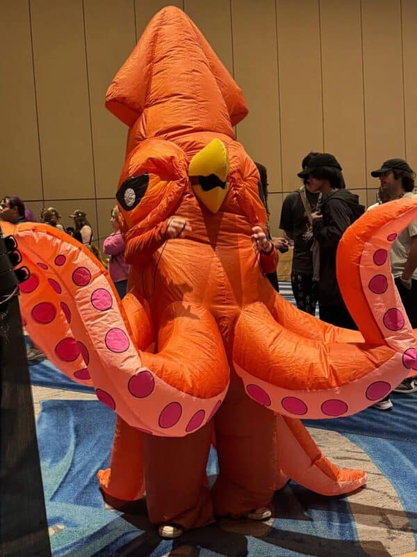 A person is wearing an inflatable, orange squid costume with large cartoonish eyes and a yellow beak. The costume’s tentacles are spread wide, showcasing pink circular patterns. The person inside is barely visible, with only their hands emerging from the sleeves. They are standing in a room with a patterned carpet and beige walls, surrounded by other people casually