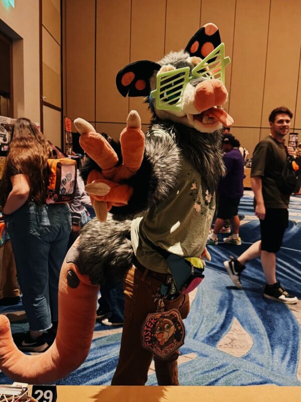 A person in a gray, furry animal costume with large ears, a snout, and a long tail poses playfully at a convention. They wear oversized green shutter shades and hold up a peace sign with their fuzzy paw. The costume has colorful accents, and a large badge with “Fusser” is attached to their belt. Other attendees are