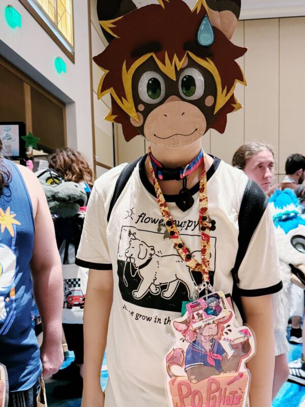 A person stands indoors wearing a colorful, animal-themed mask with large eyes, a happy expression, and a sweat drop on one side. They are dressed in a white and black T-shirt featuring a cartoon dog and text, and have a bright lanyard around their neck. Attached to the lanyard is a large, decorative badge with an illustration