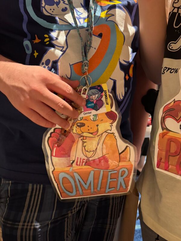 A close-up image of a person wearing a colorful graphic t-shirt with a cartoon design. They are holding a large badge featuring an illustration of a smiling cartoon animal in a lifeguard chair, with the name OMIER in bold letters. Attached to the badge is a lanyard with smaller attendee cards. Another person in a different graphic